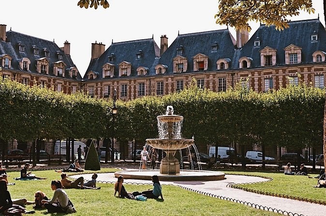 The Place des Vosges garden on a sunny afternoon, people on the lawn around a fountain and the slate roofs of the pavilions behind the lime trees