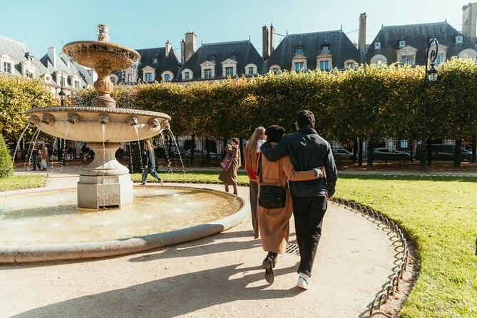 A couple walking past one of the Place des Vosges fountains, the clipped lime trees and the pavilion roofs beyond