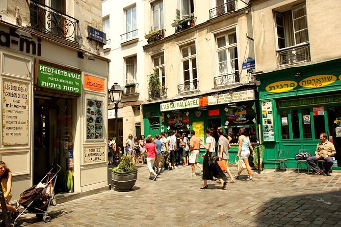 The Rue des Rosiers at lunchtime, a queue outside the falafel counters under green and yellow shop signs in French and Hebrew