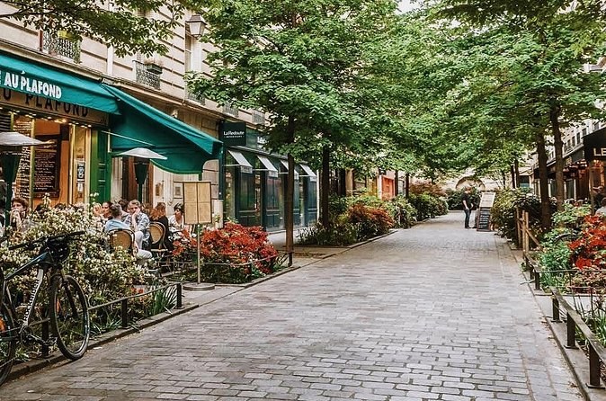 A cobbled lane under plane trees in the Marais, café terraces and flower boxes along one side